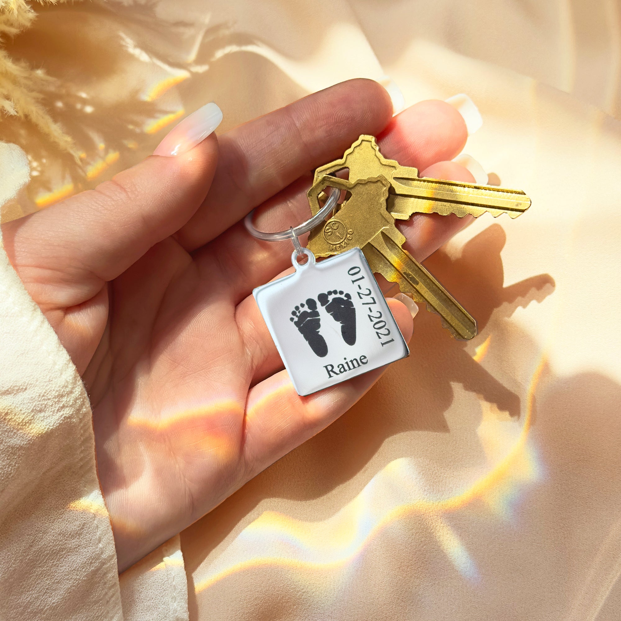 Personalized footprint keychain attached to everyday house and car keys, shown in a hand with warm sunlight to highlight the brushed metal finish and detailed baby feet engraving. Custom baby footprint keychain with name and date, a practical keepsake for new dads, moms and grandparents who want to carry their child’s first footprints wherever they go.