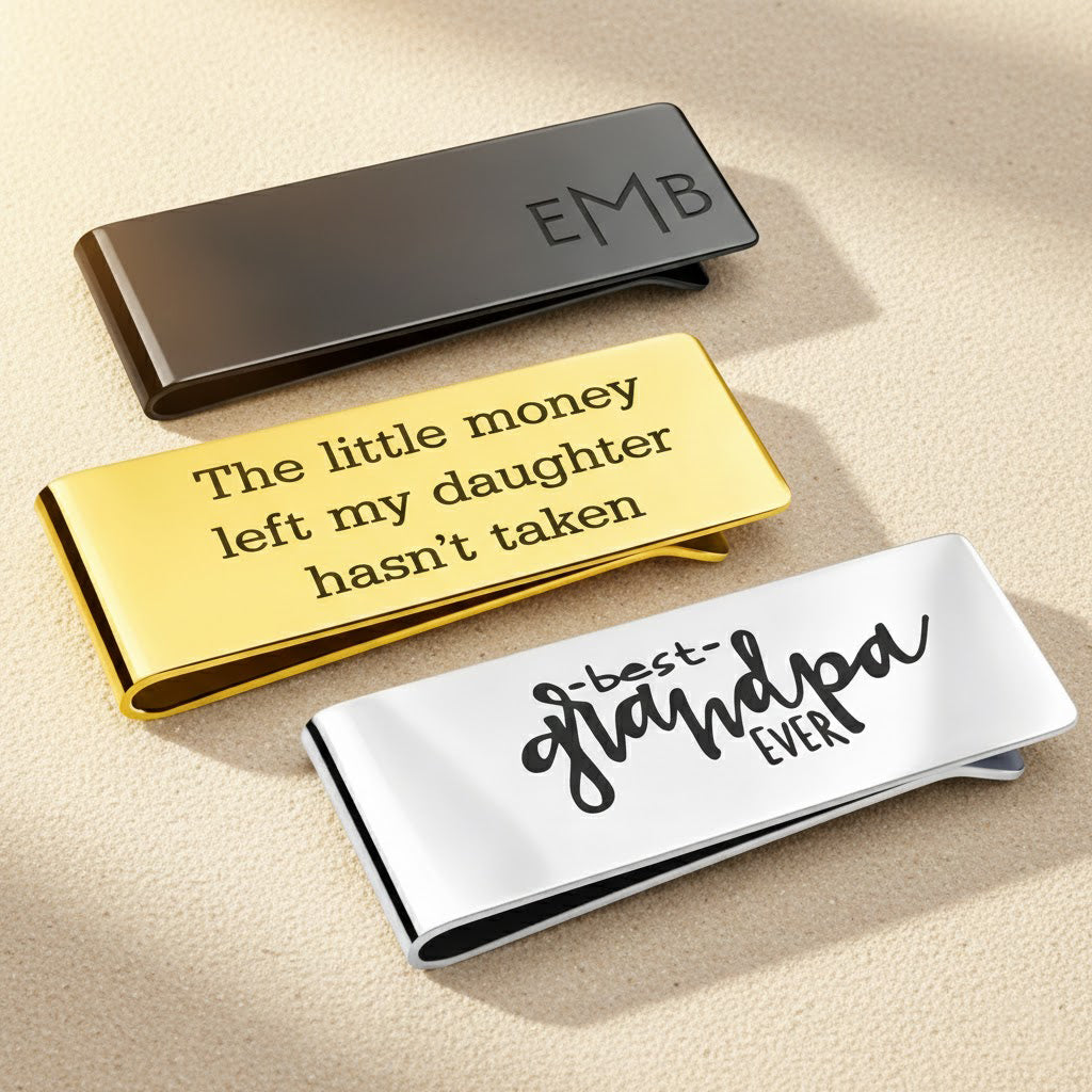 Three polished money clips in gold, black, and silver with custom engravings, displayed on a soft surface with sunlight reflections.