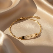 A gold bar bracelet engraved with a dog's name with a small paw print. The bracelet sits on a beige silk background.