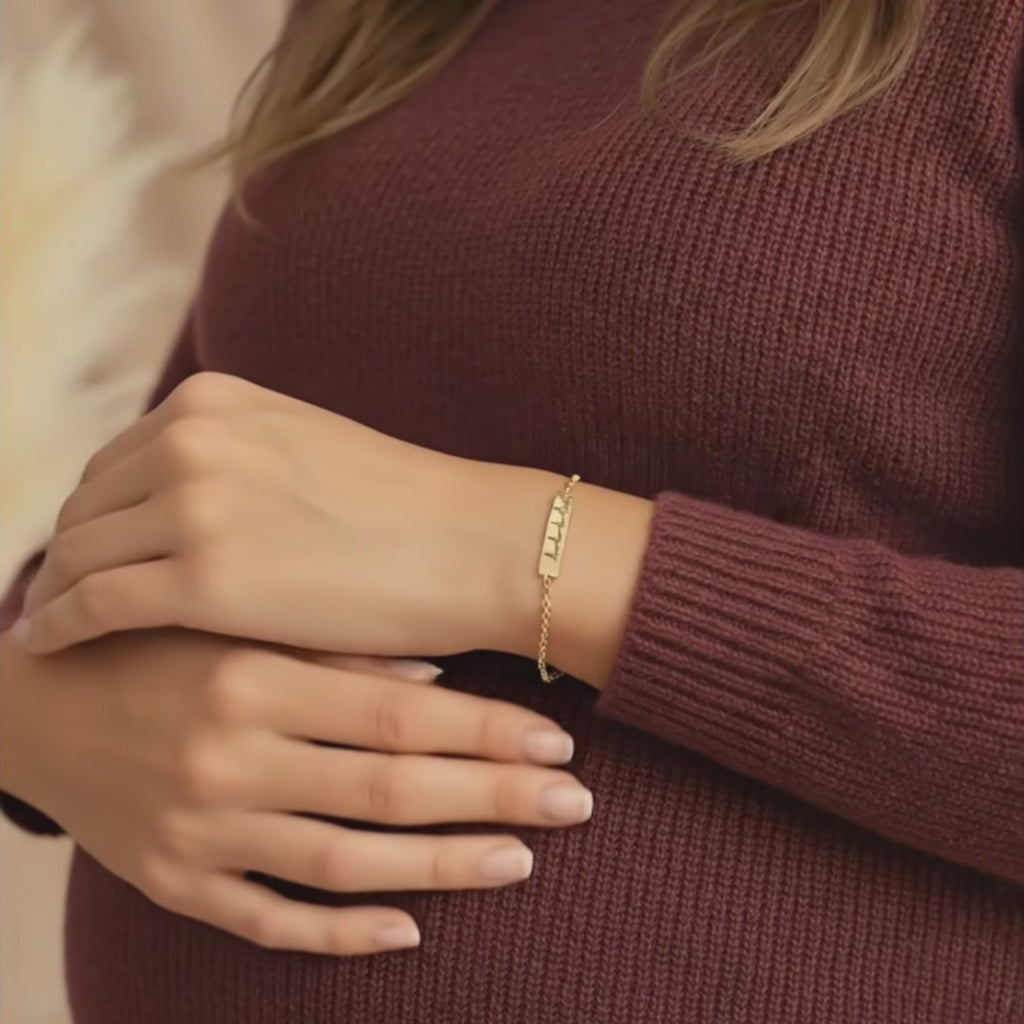 Video showcasing a pregnant mom gently resting her hand on her baby bump while wearing a custom engraved heartbeat bar bracelet in shiny yellow gold, featuring a delicate chain and minimalist heartbeat line design across the bar; sentimental maternity jewelry gift for expecting mom or new mom, perfect for baby shower, pregnancy announcement, push present or Mother’s Day, modern and elegant everyday bracelet.