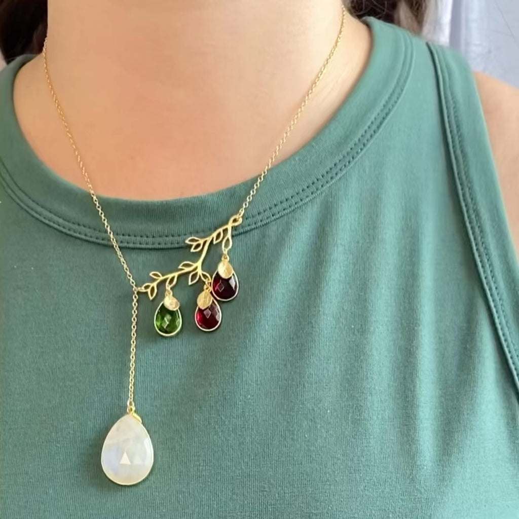 A person in a green sleeveless shirt wears a lariat branch necklace featuring a bezel set large moonstone and small peridot, small ruby and small garnet birthstone. Each stone has a dainty gold leaf initial charm overlaid on the top. Grandmother's birthstone necklace, personalized mother's and children's birthstone and initial necklaces, family branch family tree necklace, custom family tree branch necklace in gold and silver. Represent everyone in the family with their birthstone and initial charm.