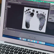 A set of baby footprints on a computer screen slides to the left to reveal a solid black set of the same footprints indicating editing. A person wears a gold footprint bangle with a yellow citrine birthstone. Two hands hold the same bracelet up close to the camera to reveal the footprints from the first shots engraved above the name Rachel 8 - 5 - 24 in Modern Thin font. A person wearing the same bangle turns their wrist to show the clear engraving. A silver bangle with a blue kyanite birthstone in box.