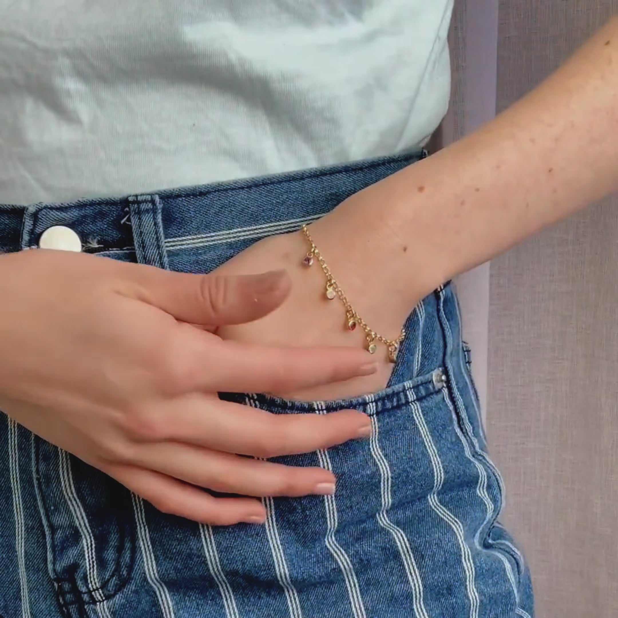 A person wears a dangling birthstone bracelet with their hand in their pocket. A close up shot of two dangling bracelets. A person wears a multiple birthstone minimalist laying bracelet and moves their arm up and down. A close up shot of a person wearing a bracelet and running their finger over the gems. Available in sterling silver, gold, or rose gold. Birthstones for every month available.