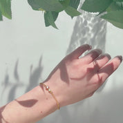 A person wears a mother and child 5mm and 3mm bar bracelet with a large Garnet and a small Aqua Chalcedony stone against a white background. Shadows and leaves can be seen in frame. A panover shows 3 gold bar bracelets with different styles of bar bracelet, each with a different array of birthstones. A persons hand shows the bracelets stones over their knuckles, shining bright in direct sunlight. A bracelet with a Garnet mother's stone and an Aqua Chalcedony child's stone is in an open Danique Jewelry box. 