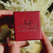 A person opens a red Danique Jewelry ring gift box to reveal a gold vermeil engraved oval birth flower ring. A person wears a silver engraved birth flower ring with a stackable citrine diamond shaped ring against a tree. A hand holds a silver engraved birth flower ring against a blue sheet background. An up close shot of a person's hand wearing a sterling silver ring. 