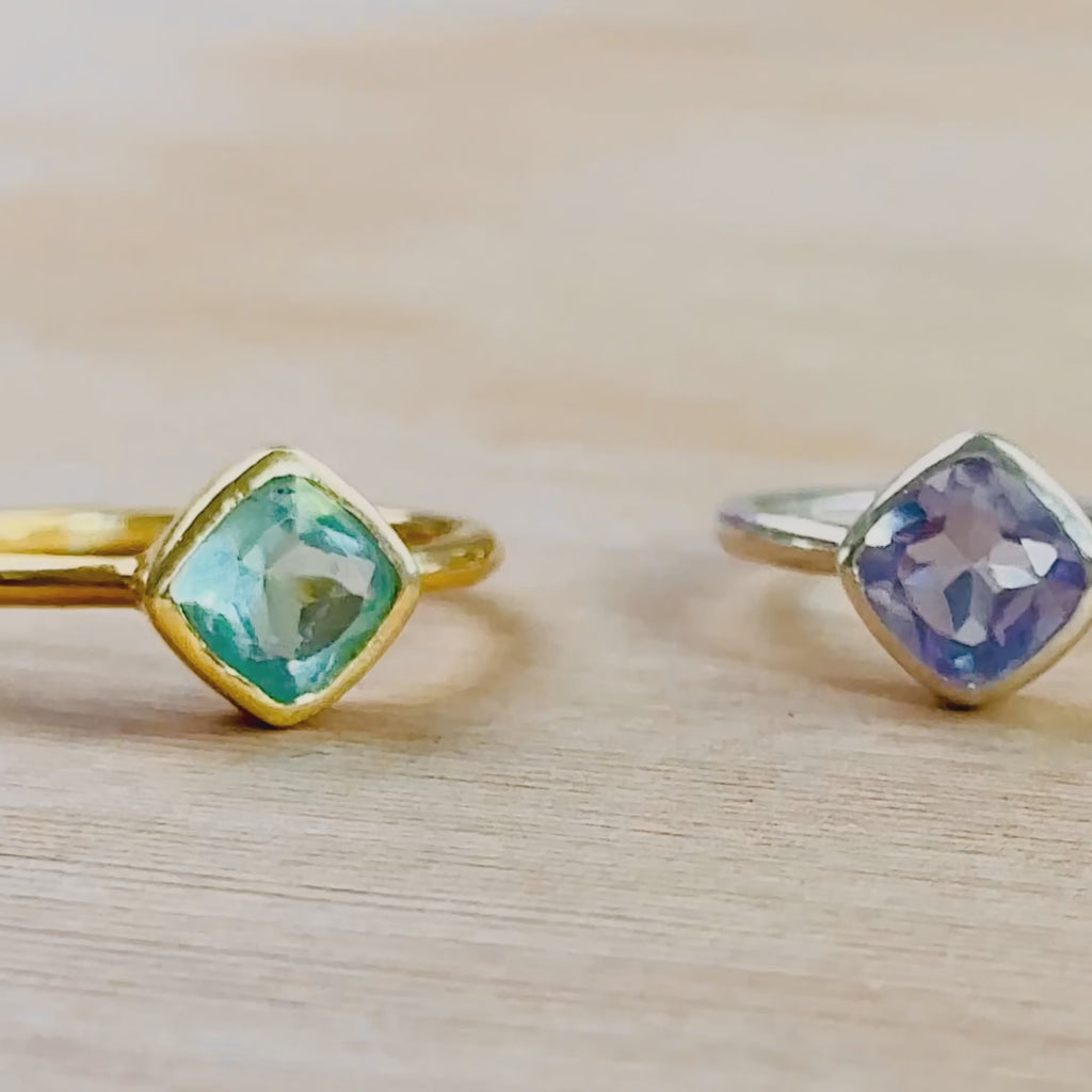 A camera pans horizontally to reveal blue topaz, alexandrite and clear quartz diamond rings. A person holds a gold set blue topaz diamond shaped ring up close. A person wears a gold moonstone ring on their pointer finger in the sunlight. A different person wears a stacked set of two rings in silver with kyanite and alexandrite. The same ring set is then shown in an open red Danique Jewelry box. Ideal for birthdays, Christmas, or as a Mother's Day gift for her.