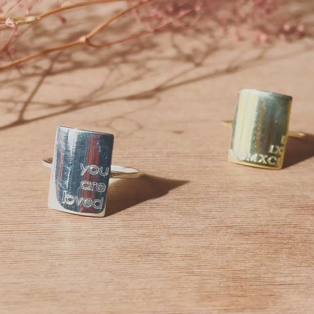 Camera pans over 3 engraved rectangle minimalist rings in silver, gold vermeil and rose gold vermeil. Each ring is engraved with a phrase or symbol. The second clip shows a rectangle ring on a hand shining in the sun engraved with the words "You are loved". A close up of the ring shows deep engraving and easy to read words. The final shot shows  a vermeil gold rectangle ring with roman numerals in an open red Danique Jewelry gift box.