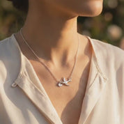Gold mama bird necklace with two tiny baby bird charms engraved with initials L and K, resting on soft ivory fabric in warm sunlight. Personalized family necklace symbolizing motherhood, children, and love.