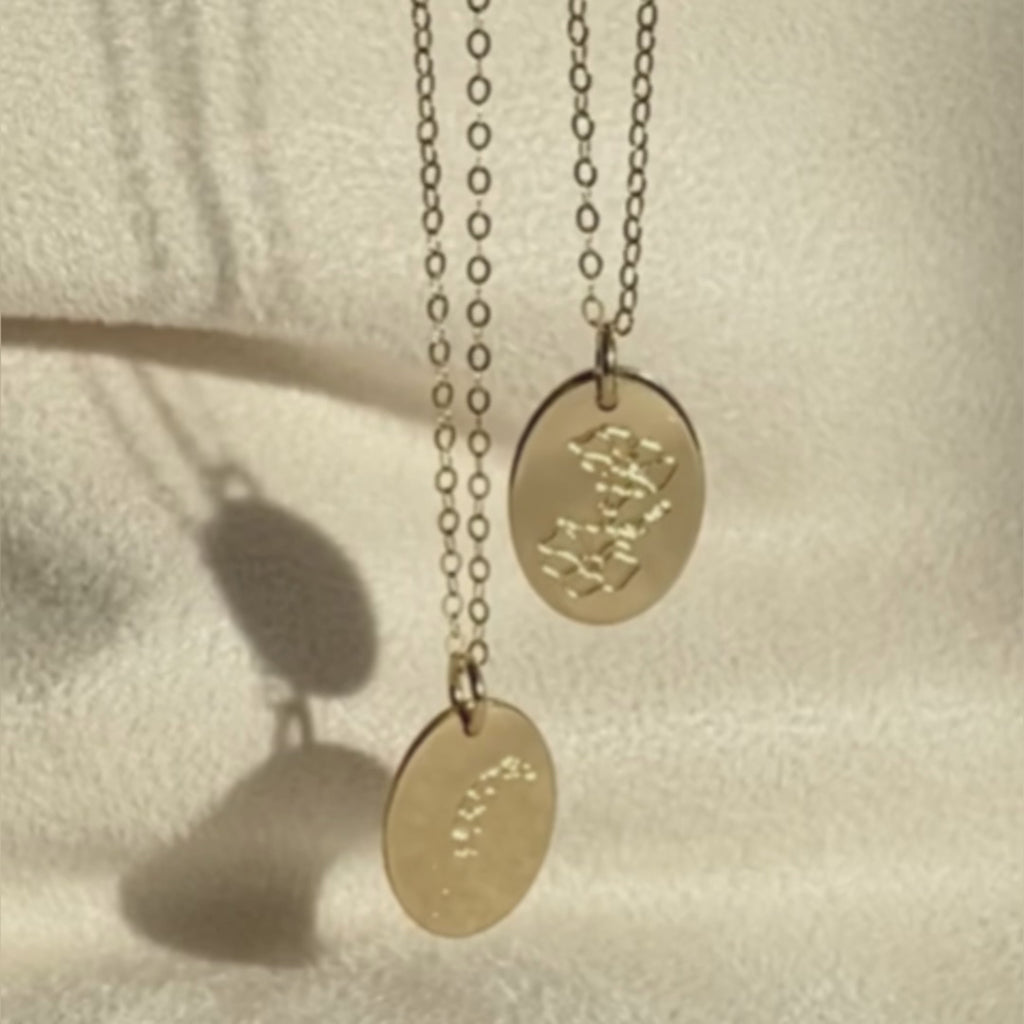 2 Engraved birth flower necklaces dangle in front of a satin background. The shot fades to a still photo of four engraved oval disc birth flower necklaces in a zig zag pattern. Text in the bottom left corner reads "find your flower" in a white cursive script.  Next to the third flower, "May" appears, followed by the first flower showing "July" on the side. The fourth necklace reads "August" next to the piece, and the second necklace shows "September" in the same font. Personalize with your birth flower!