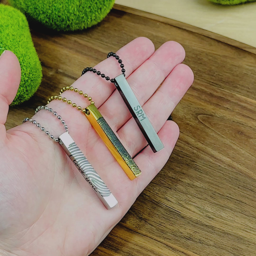 A video shows off an assortment of 3D Bar men's necklaces in silver, gold and black stainless steel. 3 necklaces rest in a person's hand before shifting to a close up of a silver necklace on a man's chest. The bar can be engraved with fingerprints, handwriting, text, and small simple images. A person shows a picture of a fingerprint next to the same fingerprint engraved on a silver bar necklace. The video finishes with a gold fingerprint necklace in a box with the side engraved which says "my love".