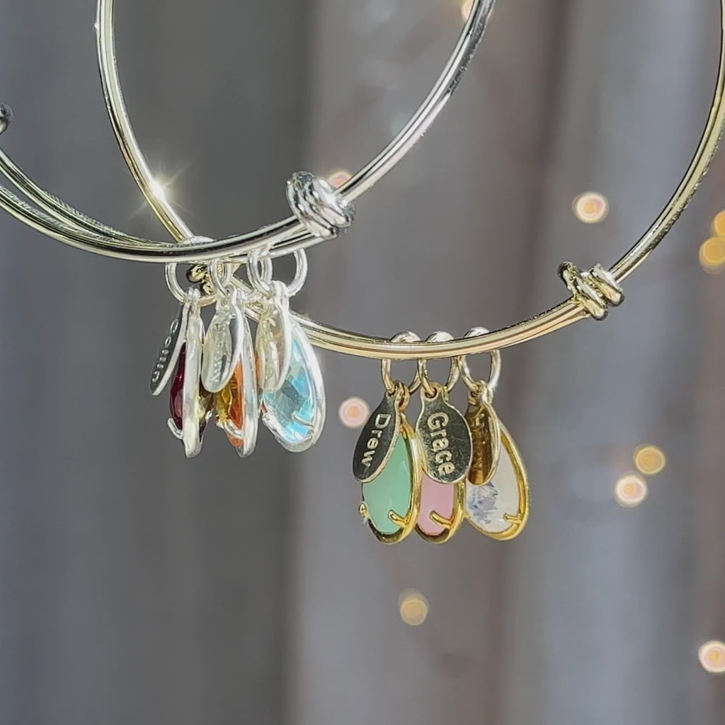 A gold and a silver bangle bracelet with engraved name tag charms and half bezel prong teardrop birthstones. A person wears a gold bangle bracelet with three kids names and birthstones. A silver bangle bracelet in an open red Danique Jewelry box. Personalized bracelet for mom with kids names and gemstones, multi stone multi color charm bracelet bangle style for mother for women