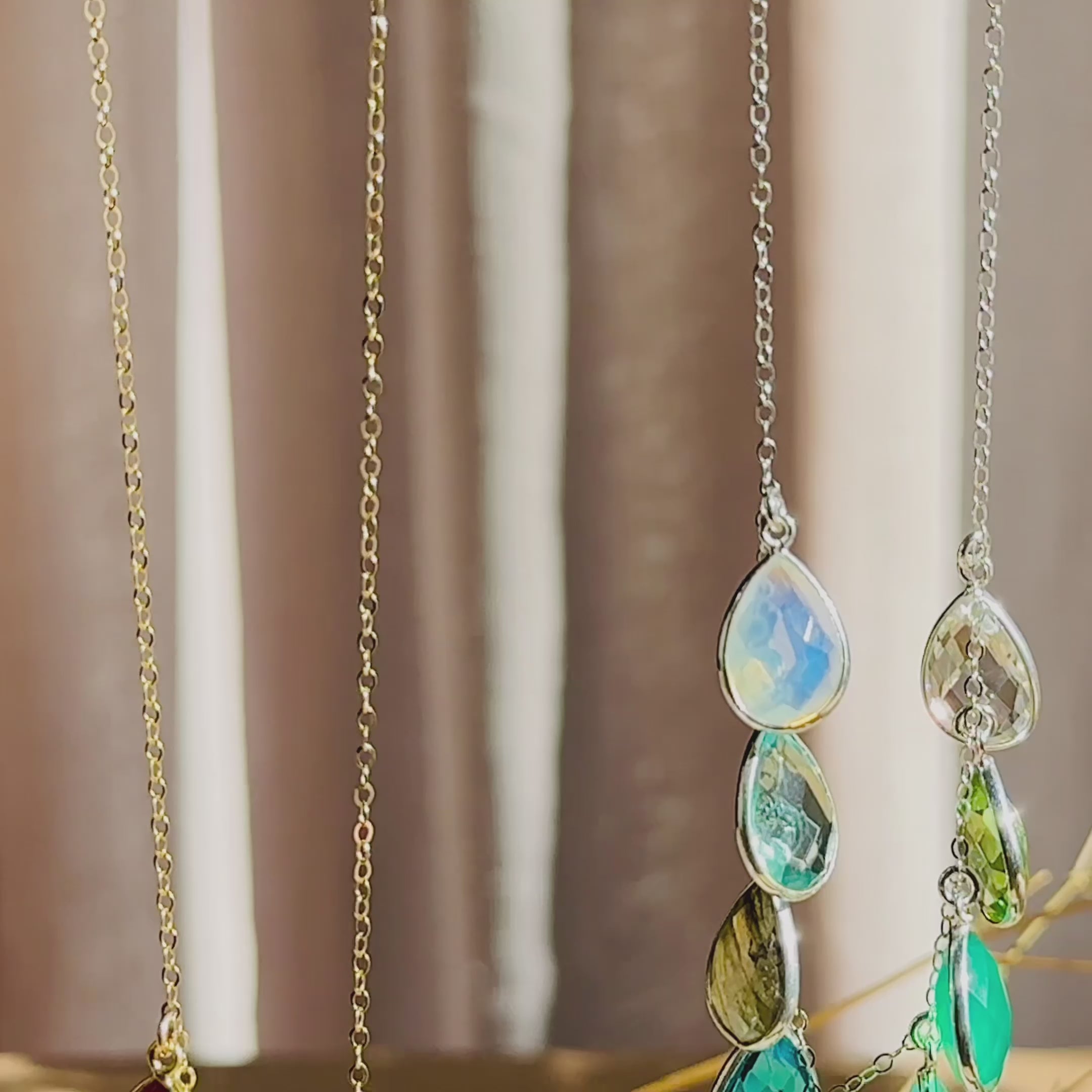 The camera pans down over a silver and a gold birthstone collar necklace. A person in a black shirt wears a multi stone teardrop collar necklace. Two hands gently tilt and shift a necklace back and forth to show the movement of the birthstones. A person opens a red danique jewelry gift box to reveal a silver teardrop necklace with multiple birthstones in varying shades of green and blue. Perfect for birthdays, christmas, mother's day, anniversaries or any special gift giving occasion for mom, grandma & wife