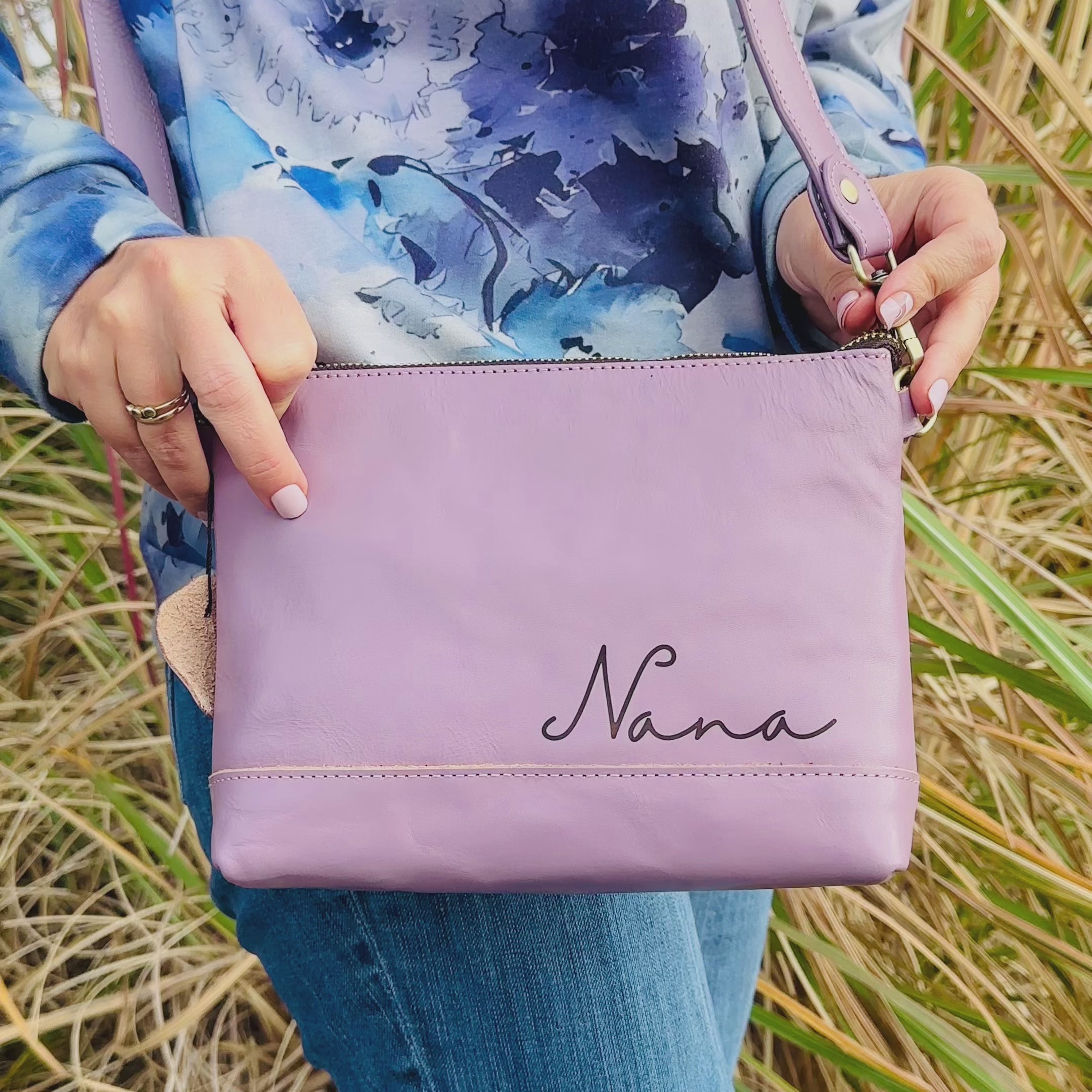 A person holds a lavender engraved leather crossbody convertible bag monogrammed with Nana. A woman holds a raspberry colored bag in front of her and shows off a monogram engraving on the front. She expertly unclasps the long strap on the bag and replaces it with a shorter clutch strap using simple clasps on either side of the bag. She turns to show the bag in full effect. The video ends with the woman holding 2 styles of engraved women's bags - a crossbody with a strap and a handheld cosmetic travel bag. 