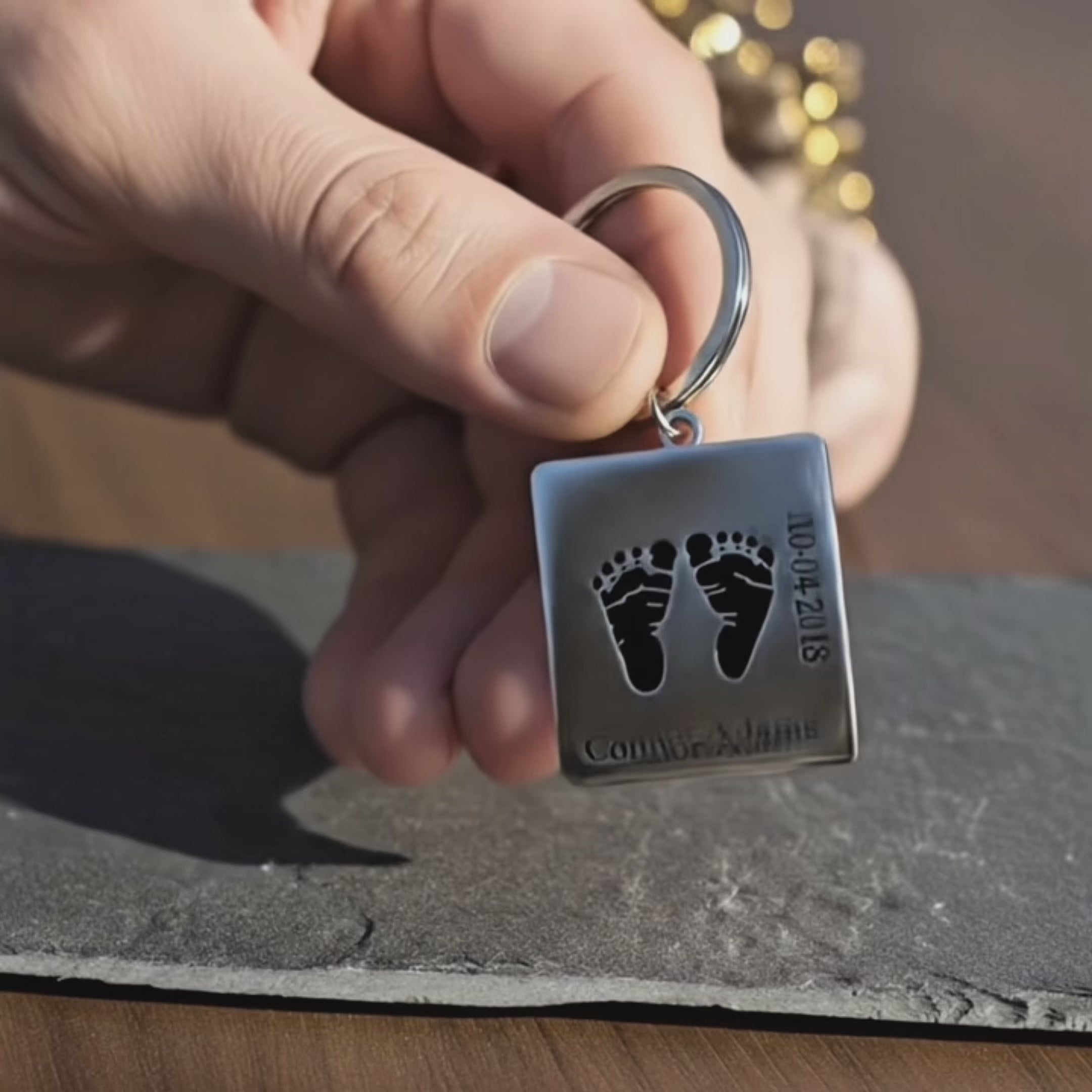 Custom engraved footprint keychain on a dark stone surface, spotlighting the crisp black baby feet artwork and personalized name on the stainless steel tag, used as the cover frame for a product video. Modern, gender-neutral new baby gift or memorial keychain that keeps your child’s footprints close, ideal for Father’s Day, Mother’s Day or remembrance gifts.