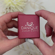 Two hands open a red Danique Jewelry ring box revealing a silver engraved coordinates thin stacking ring. A person wearing a pinky ring slides a matching ring engraved with initials onto their partner's pinky. The partner slides the pinky ring onto their femme partner's hand. A femme person crosses pinky with their partner's pinky. The camera pans over a collection of engraved rings in open red Danique Jewelry boxes. Sterling Silver, Gold Vermeil, Rose Gold Vermeil, Anniversary rings, custom wedding rings