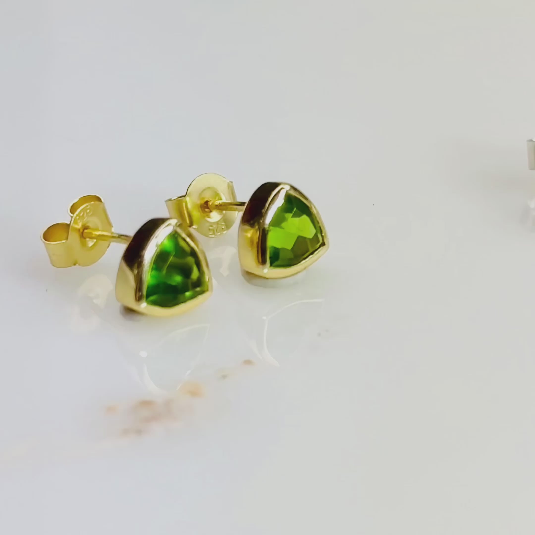 The camera pans over an assortment of silver and gold dainty triangle gemstone birthstone earrings. A hand holds and gently rolls a pair of peridot triangle studs back and forth. A person opens a red Danique Jewelry box revealing a set of purple amethyst silver triangle post earrings. A person pushes their hair back behind their ear and reveals a gold peridot birthstone stud earring. Perfect gift for birthdays, Christmas, Mother's Day, anniversaries or any special occasion. 18k Vermeil Gold or 925 Silver.