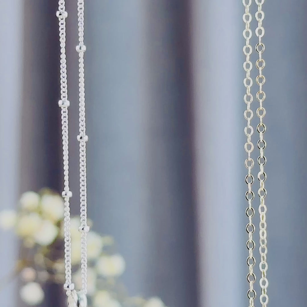 The camera pans down to reveal two round cascade necklaces in silver and gold. A person in a white shirt with long brown hairs wears a gold round cascade necklace with citrine quartz and ruby quartz suspended on a gold filled cable chain. The person thumbs a 2-stone necklace, showing off the faceted sparkling birthstones. A person shows off a three stone silver cascade necklace with blue and green stones in an open red danique jewelry box. 