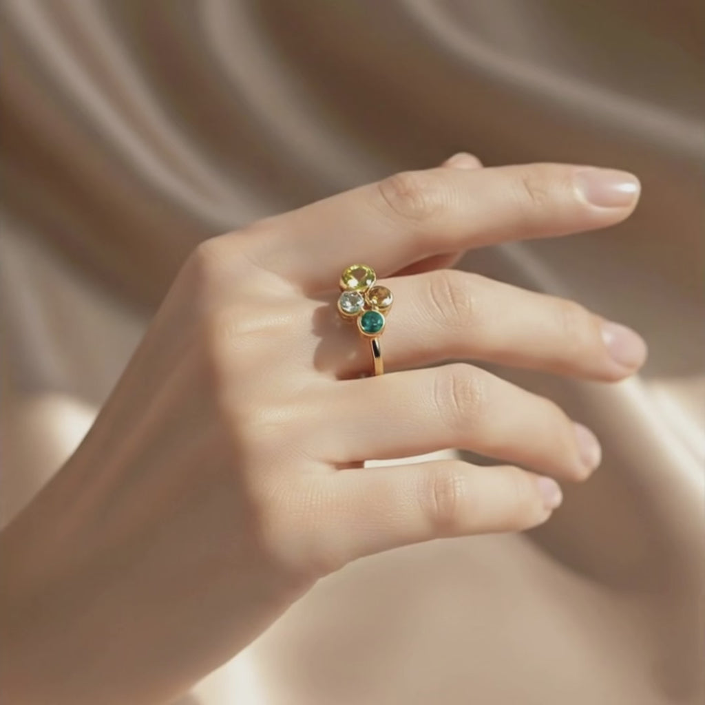 Video showing a close-up of a woman’s hands gently turning a custom birthstone ring for mom, featuring a yellow gold band with four bezel-set multi-colored birthstones symbolizing children or family, sparkling against a soft satin beige background; personalized family ring, dainty stacking style, meaningful Mother’s Day or birthday gift for mom or grandma.