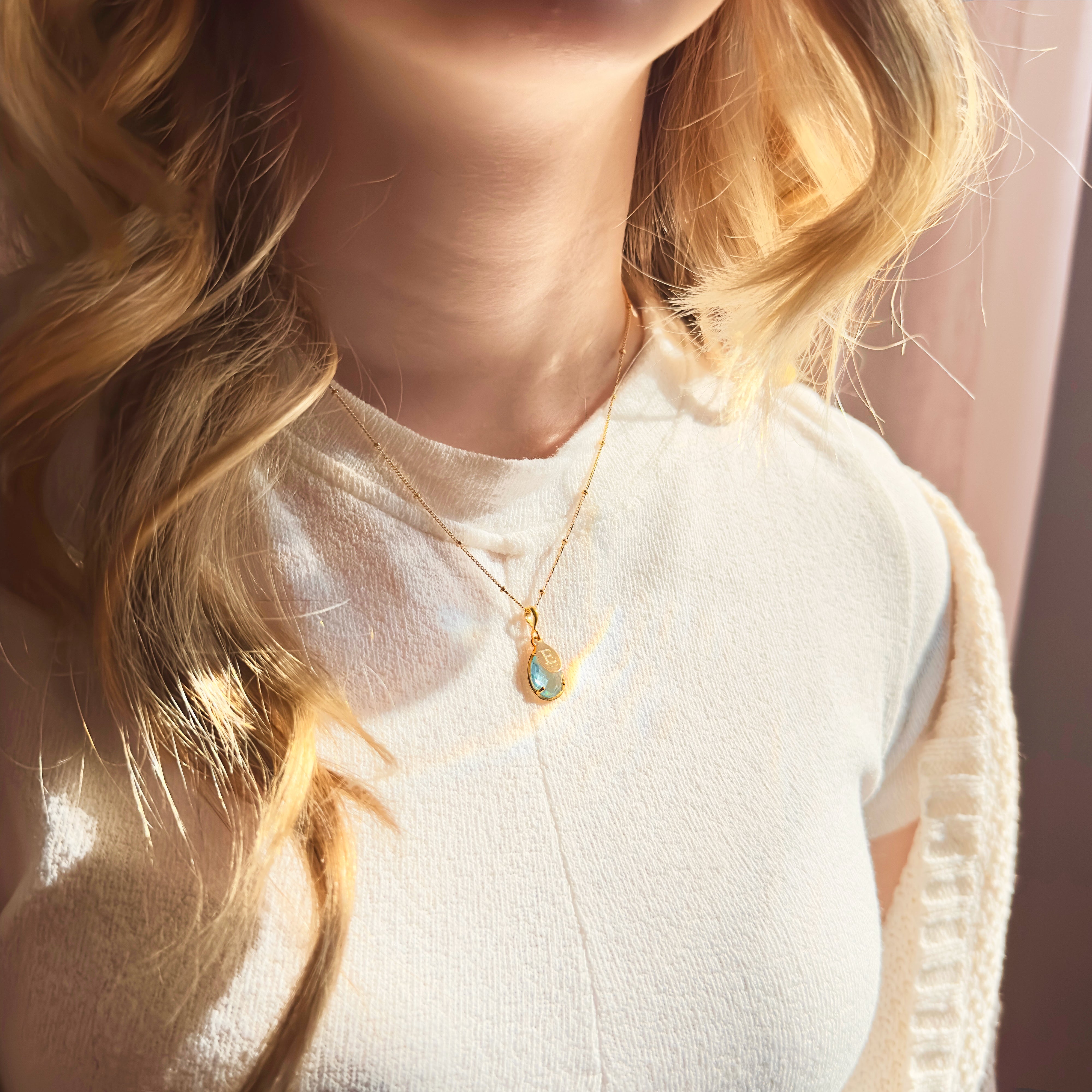 Model in beige top is wearing a prong teardrop blue topaz birthstone necklace with a classic initial engraved. December birthstone, december jewelry, blue stone, blue birthstone