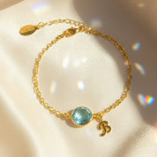 Gold personalized birthstone initial bracelet displayed on satin fabric with shimmering light reflections, featuring a bezel-set aqua blue round gemstone and matching gold script initial charm. Crafted in 18K gold vermeil with adjustable chain length, this custom birthstone bracelet makes a meaningful jewelry gift for mom, daughter, sister or best friend. Ideal for birthdays, Mother’s Day, anniversaries, Christmas or as a sentimental everyday bracelet.