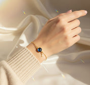 Dainty personalized birthstone initial bracelet on a woman’s wrist, featuring a deep blue faceted round gemstone bezel-set in warm gold with a tiny matching script initial charm hanging beside it on a delicate chain. Styled with a cozy neutral sweater against a soft satin background with rainbow light reflections, this custom birthstone initial bracelet is a meaningful gift for mom, daughter or best friend and perfect for birthdays, Mother’s Day, Christmas or everyday wear.