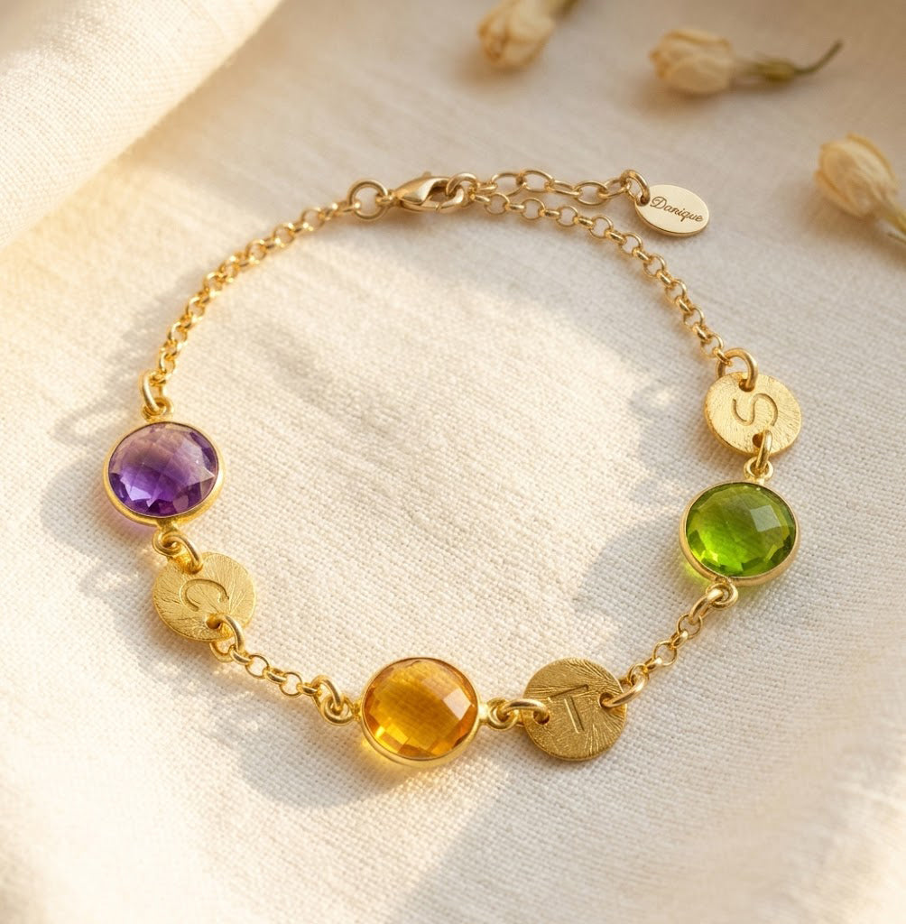 A gold bracelet featuring peridot and citrine and purple amethyst birthstones rests on a white background. Each birthstone is accompanied by a round disc charm with an initial hand stamped into it. August birthstone and September birthstone, green gemstone, purple gemstone, yellow gemstone, custom made personalized mother's bracelet with birthstones