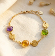 A gold bracelet featuring peridot and citrine and purple amethyst birthstones rests on a white background. Each birthstone is accompanied by a round disc charm with an initial hand stamped into it. August birthstone and September birthstone, green gemstone, purple gemstone, yellow gemstone, custom made personalized mother's bracelet with birthstones