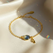 Close-up of a personalized birthstone initial bracelet featuring a labradorite bezel-set gemstone with iridescent blue-green tones paired with a dainty gold script initial charm. Set on an 18K gold vermeil chain with extender, this custom bracelet offers modern minimalist elegance. A beautiful personalized jewelry gift for mom, daughter or best friend, perfect for birthdays, Christmas, Mother’s Day or everyday stacking with other fine gold bracelets.