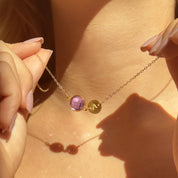 Dainty custom initial birthstone necklace in warm sunlight, featuring a faceted round purple Amethyst bezel-set in gold next to a small engraved initial disc on a delicate gold chain. Personalized minimalist jewelry perfect for mom, daughter or best friend, February birthday or everyday layering.
