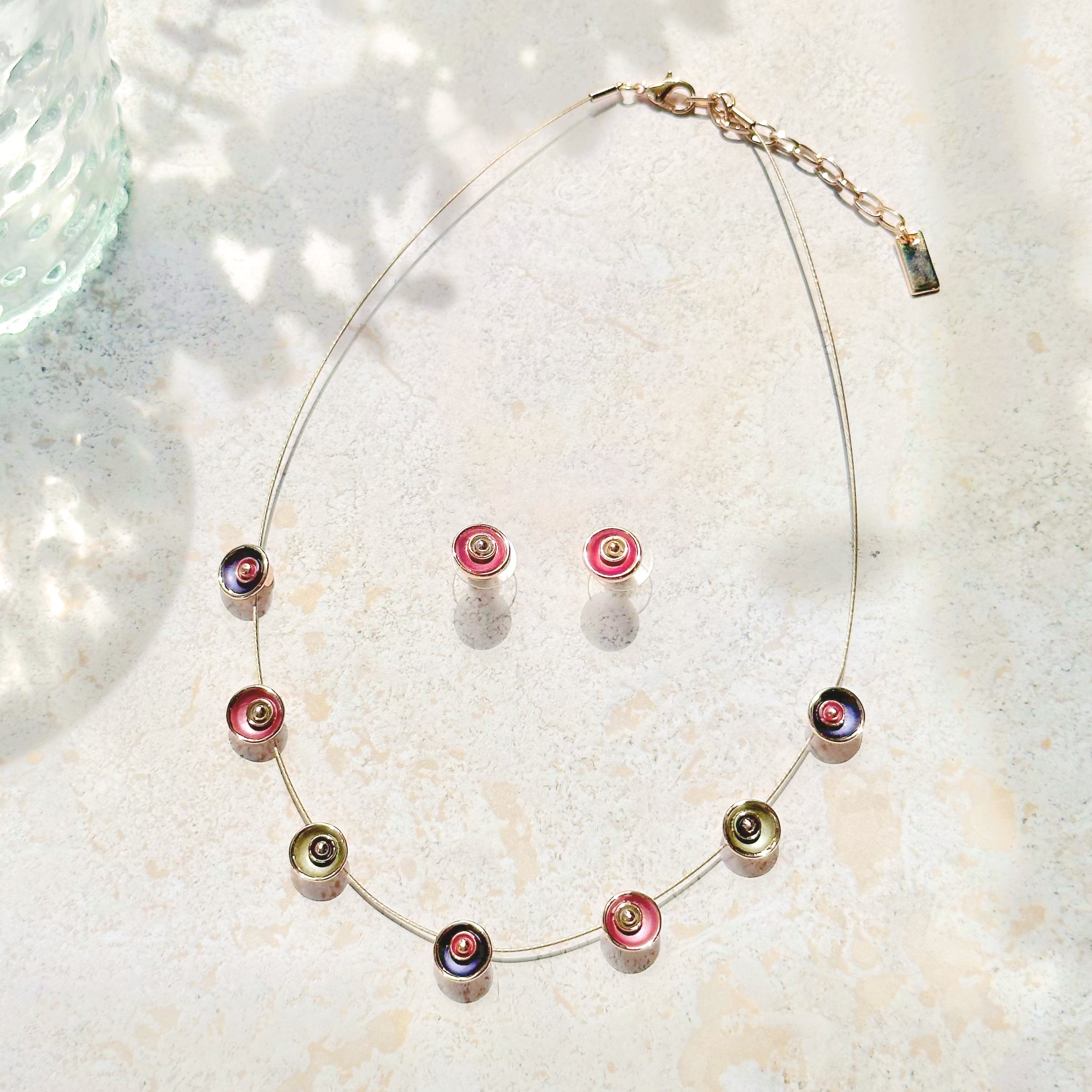 Adjustable Choker Necklace against a textured stone background with a brown steel cable chain and seven rose gold plated circular flower charms painted in purple, pink, and gray with a pair of matching circular flower earrings in pink and gray