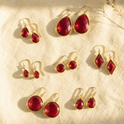 Ruby quartz earrings collection with multiple July birthstone earring styles shown on cream fabric in warm natural sunlight.