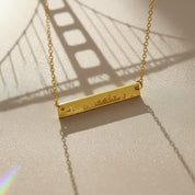 Engraved city skyline bar necklace photographed with Golden Gate Bridge shadow, personalized travel lovers necklace in gold.