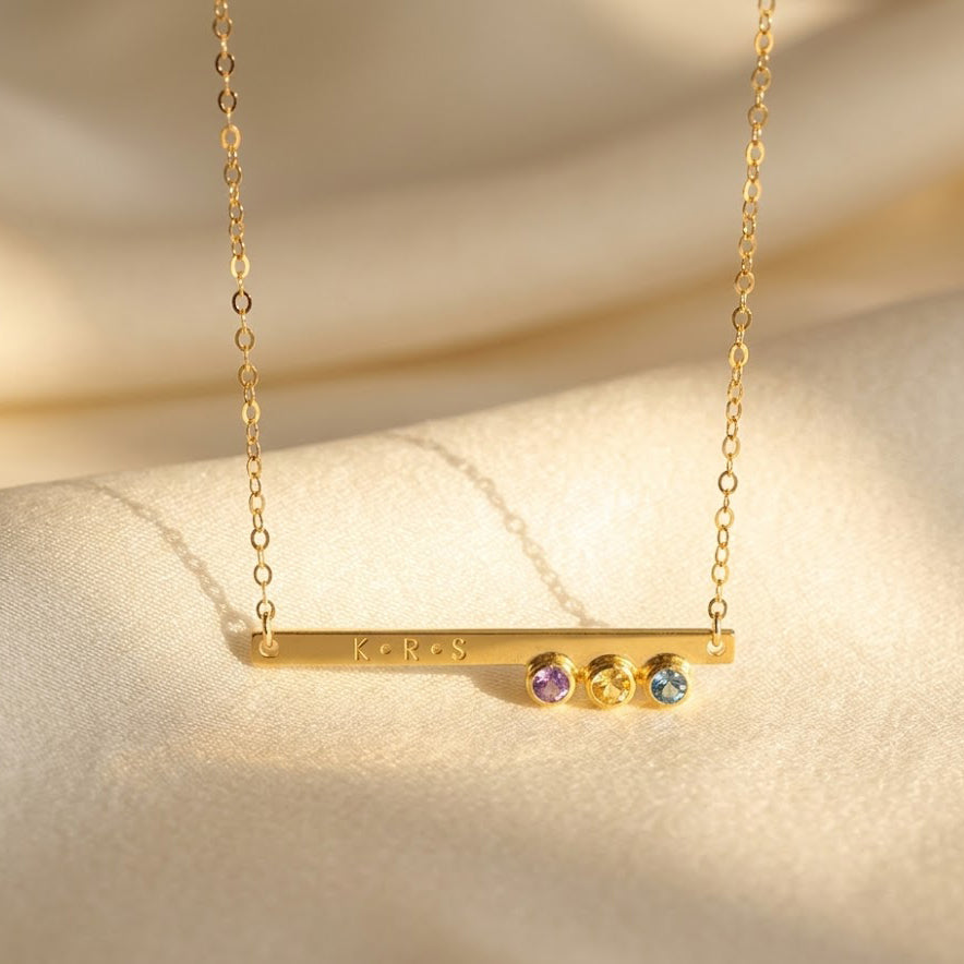 Dainty gold birthstone bar necklace with engraved name and tiny gemstone charms on beige fabric