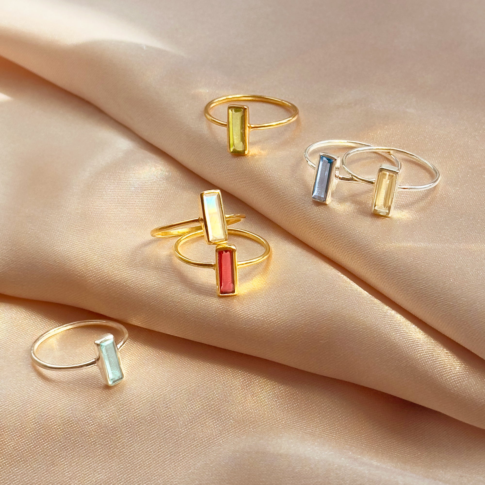 Custom birthstone baguette rings scattered on soft champagne satin, featuring slim vertical bar gemstones in pastel yellow, blue, mint, opal white and coral set in delicate .925 sterling silver and 18K vermeil gold bands. Modern minimalist stacking rings that can be personalized with your birthstone, perfect gift for birthday, anniversary, bridesmaids or Mother’s Day.