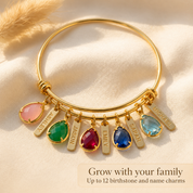Gold personalized family bangle with five colorful teardrop birthstones and engraved name charms on cream fabric.