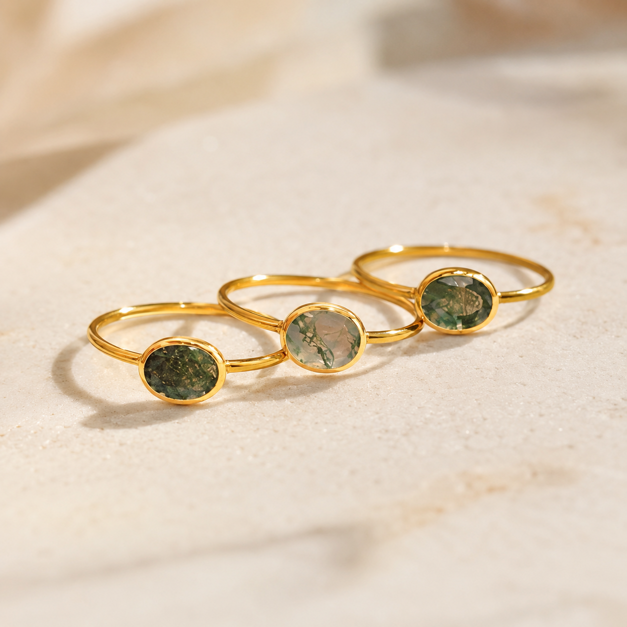 Three gold rings with oval green stone inlays, arranged diagonally on a light beige textured surface.