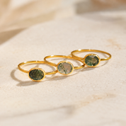 Three gold rings with oval green stone inlays, arranged diagonally on a light beige textured surface.