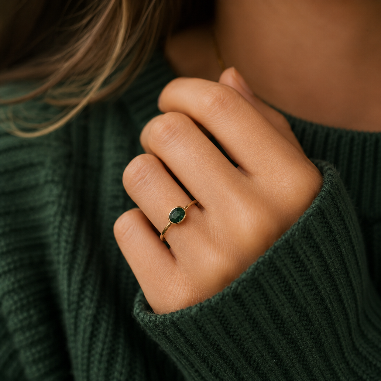 A close-up of a person's hand wearing a delicate gold ring with a small oval green gemstone, styled with a dark green ribbed knit sweater.