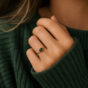 A close-up of a person's hand wearing a delicate gold ring with a small oval green gemstone, styled with a dark green ribbed knit sweater.