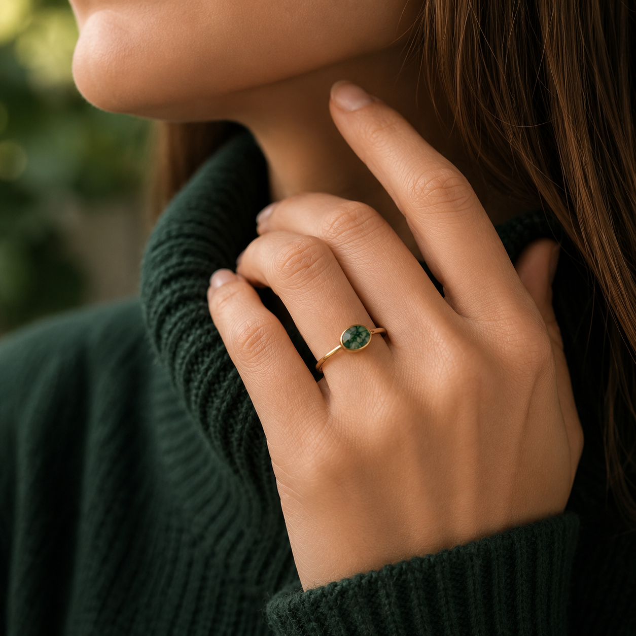 A close-up lifestyle shot of a woman wearing a delicate gold ring with a round green gemstone on her ring finger, paired with a dark green ribbed knit turtleneck sweater.