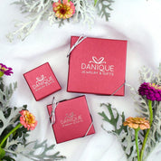 Three Danique Jewelry Branded Red Pink Gift Boxes for Ring, Necklace, and large Necklace with silver bows on a white cloth background with flowers around them. Ready for Gifting.