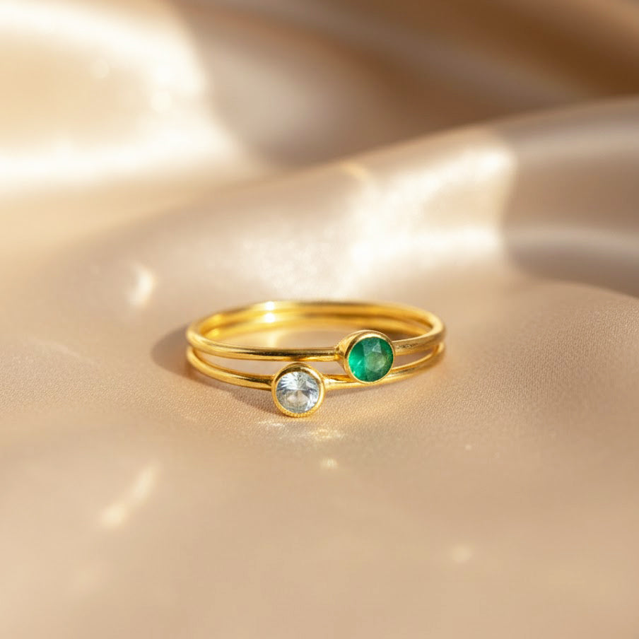pair of dainty stackable birthstone rings with slim gold bands resting on softly draped champagne satin, one bezel set with a pale icy aqua stone and the other with a rich emerald green gemstone, showcasing the minimalist design of these thin aqua chalcedony and green birthstone rings, perfect for creating a personalized march birthday stack or meaningful mom ring set, handmade in sterling silver or gold vermeil for everyday wear or gifting