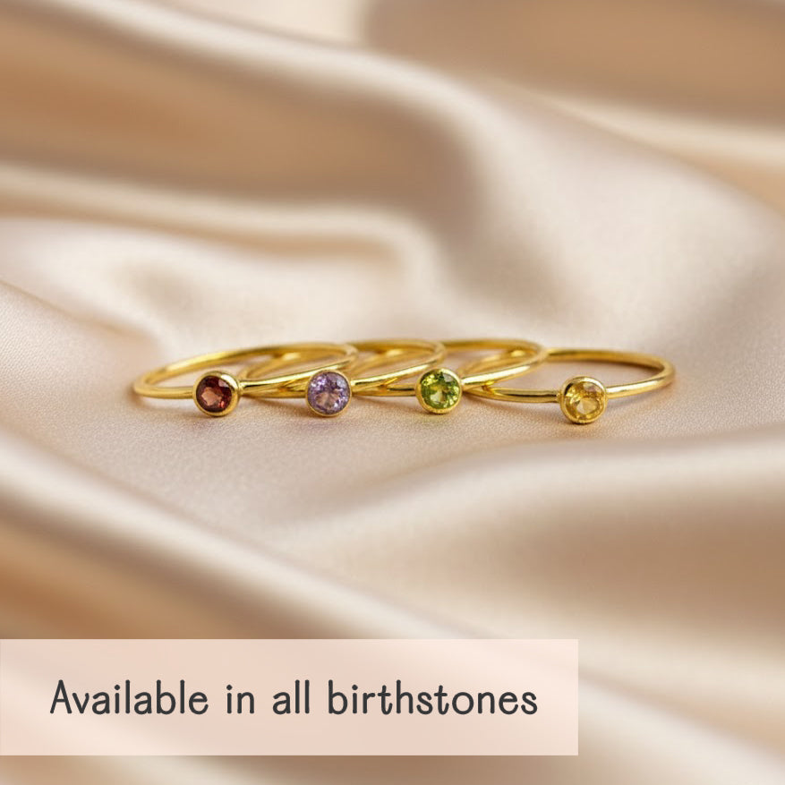 set of dainty stackable birthstone rings with slim gold bands and small bezel set gemstones in warm red, soft lavender, fresh peridot green and golden yellow, arranged in a row on champagne satin fabric with gentle folds, showcasing the minimalist design and delicate sparkle, caption reading “available in all birthstones,” perfect fine jewelry gift idea for creating custom birthstone ring stacks for mom, daughters, sisters or best friends