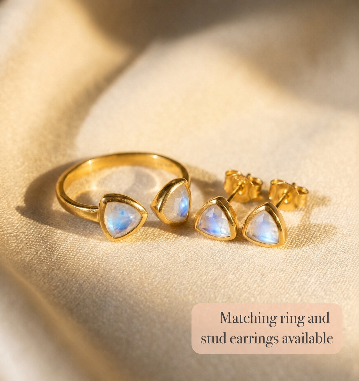 Gold moonstone triangle ring with matching stud earrings shown on warm fabric.