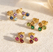 Custom triple birthstone stud earrings with three 3mm gemstones in gold settings on a soft neutral fabric background.