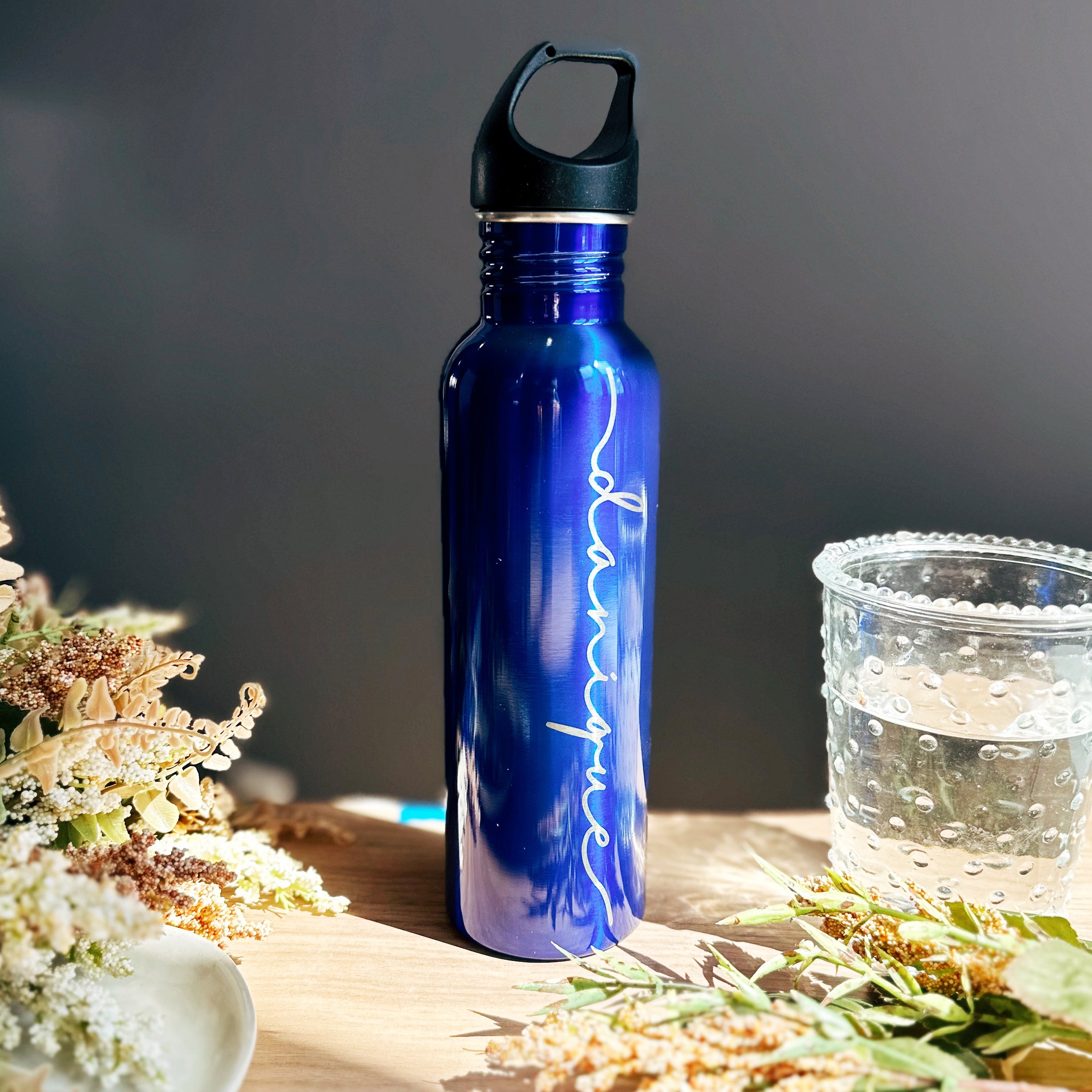 A blue water bottle with the name danique engraved vertically down the side of the bottle sits on a wood table with flowers and a glass. Personalized water bottle for gymnast, ballet water bottle personalized, travel water bottle