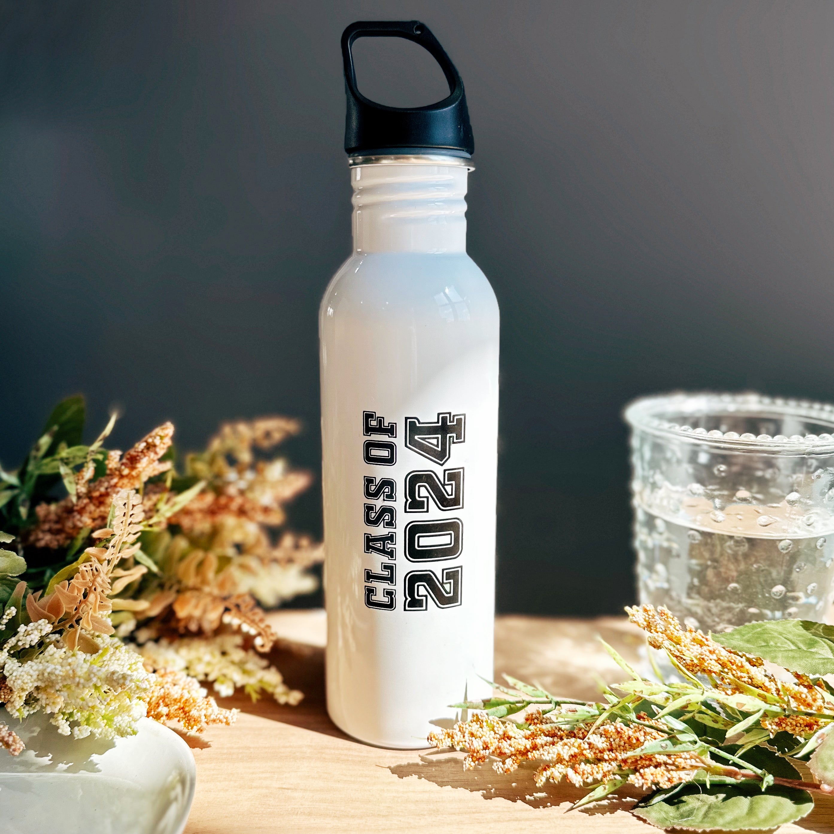 A white steel bottle engraved with the words Class of 2024 sits on a wood table with flowers and a glass. The personalized water bottle for runners, athletes, office workers, and graduates holds 26 fl oz and is great for daily use.