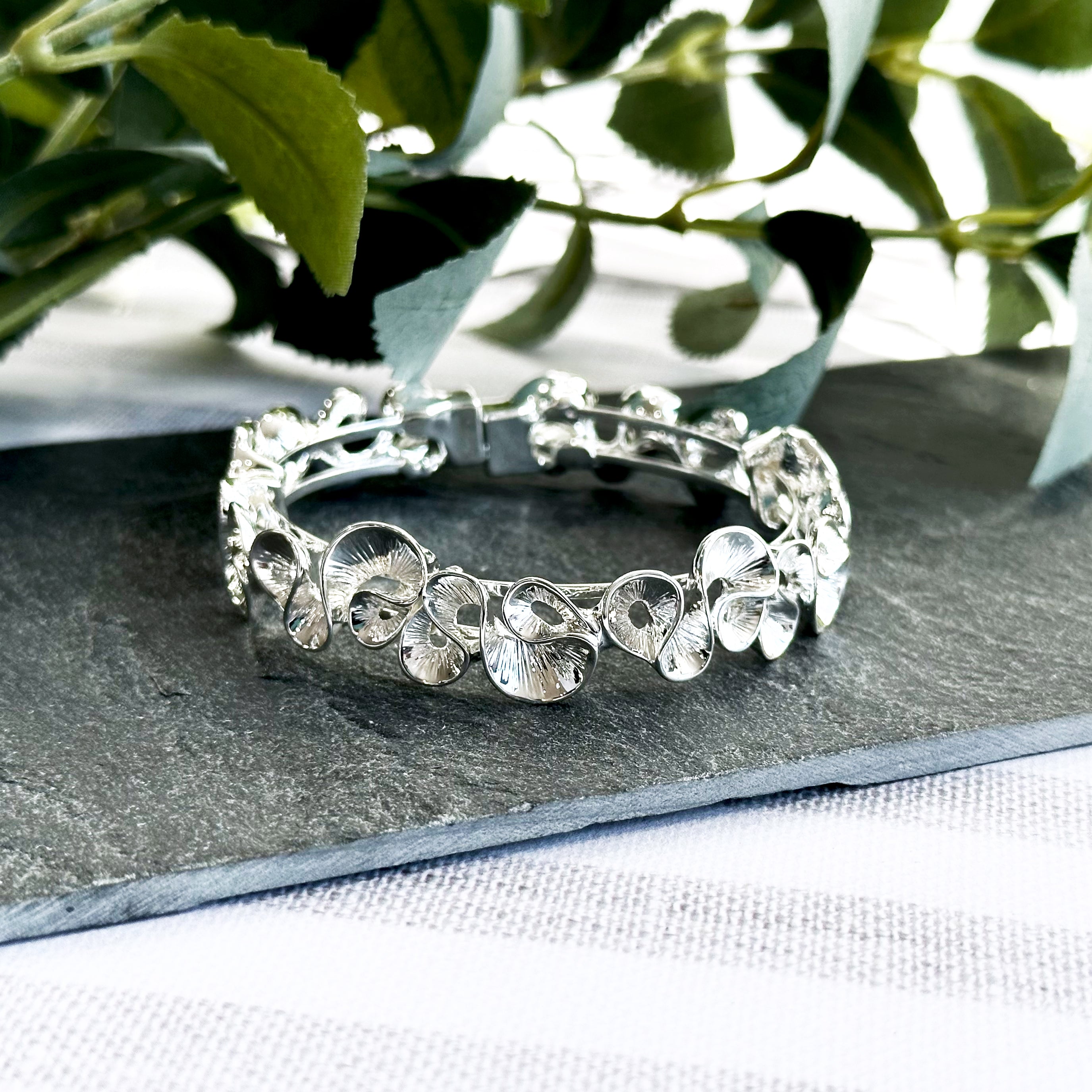 Silver Boho Style Wave Bangle Bracelet with magnetic clasp rests on a dark flat stone with leaves in the background