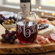 Personalized stemless wine glass with elegant engraved three-letter monogram filled with red wine, styled on a wooden charcuterie board with grapes, cheese, meats and olives; custom engraved wine glass gift for her, bridal shower, housewarming or anniversary.