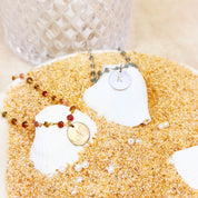 Custom initial disc pendant necklaces displayed on gemstone wire-wrapped chains with vibrant beads, styled in a beach-inspired flat lay. Personalized engraved letter charms make thoughtful gifts for bridesmaids, moms, sisters, or best friends. Elegant minimalist design perfect for birthdays, vacations, anniversaries, and everyday wear.